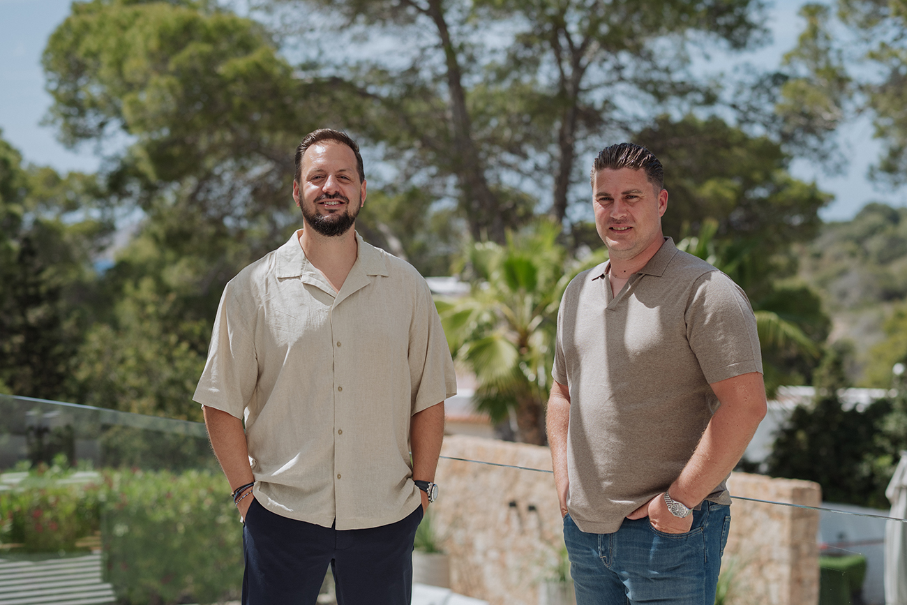 Two men standing outdoors on a balcony with greenery in the background
