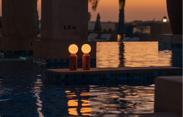 Two portable garden lamps illuminating a swimming pool at sunset with palm trees in the background.